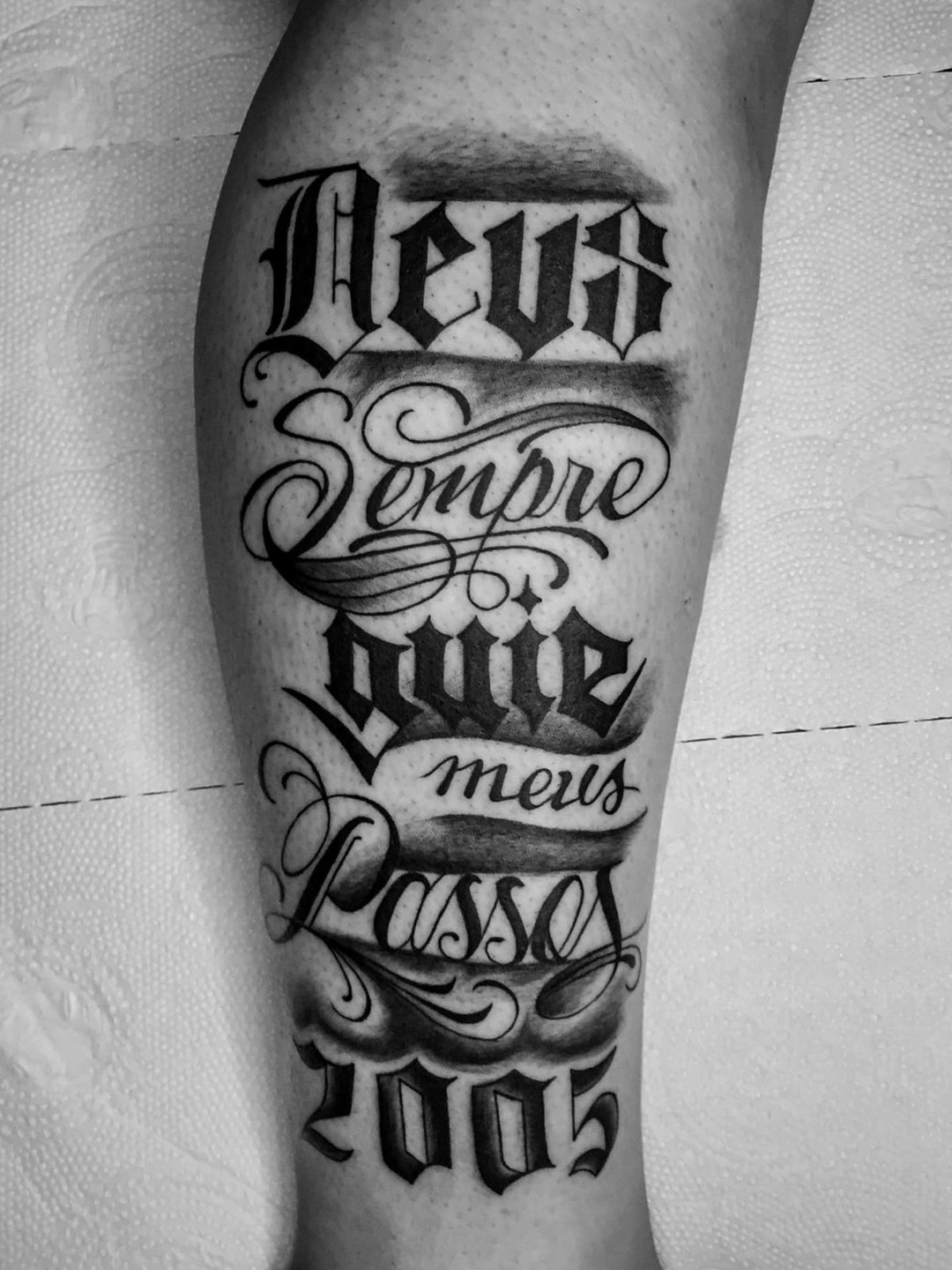 Tatuagem de lettering na panturrilha: 'Deus sempre guie meus passos 2005' em letra gótica e cursiva sombreada