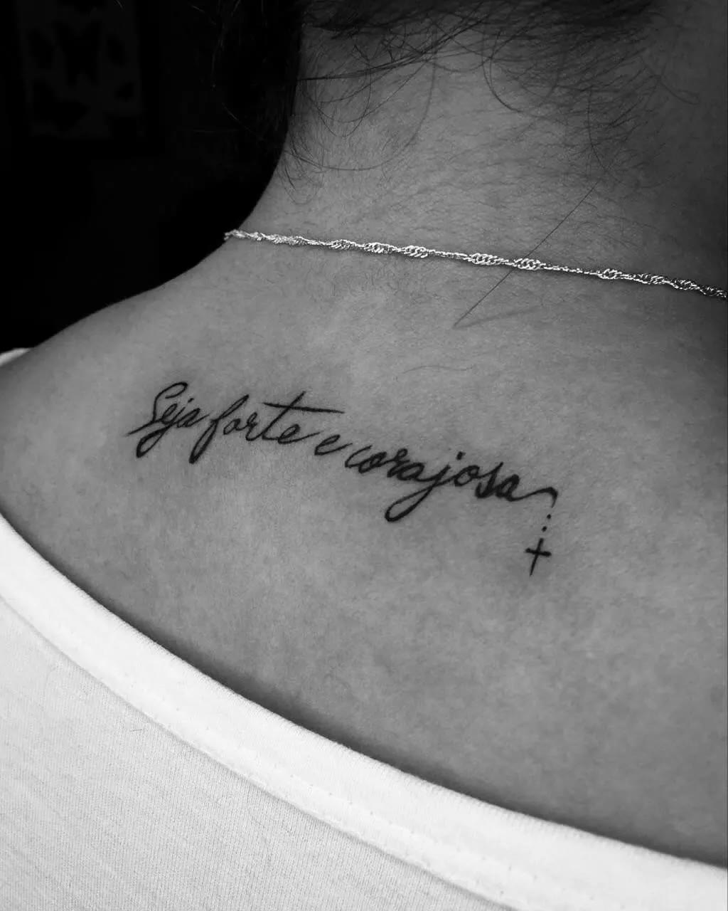 Tatuagem de lettering fine line na parte de cima das costas: 'Seja forte e corajosa' com uma cruz