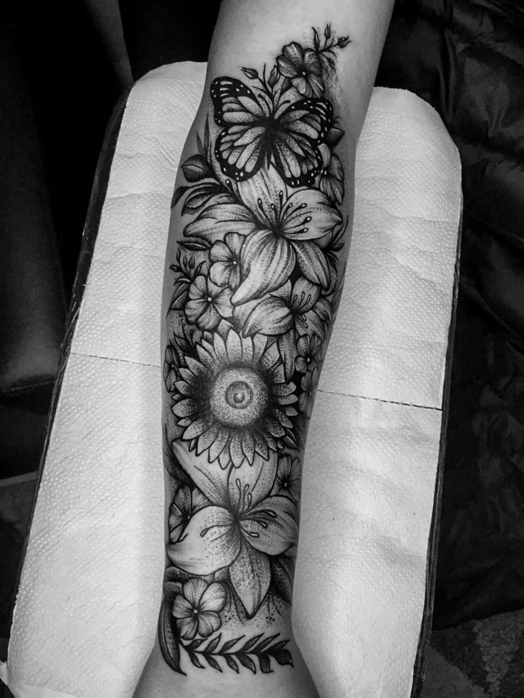 Tatuagem em pontilhismo no antebraço: arranjo botânico com borboleta, girassol, lírios e flores do campo, em preto e cinza