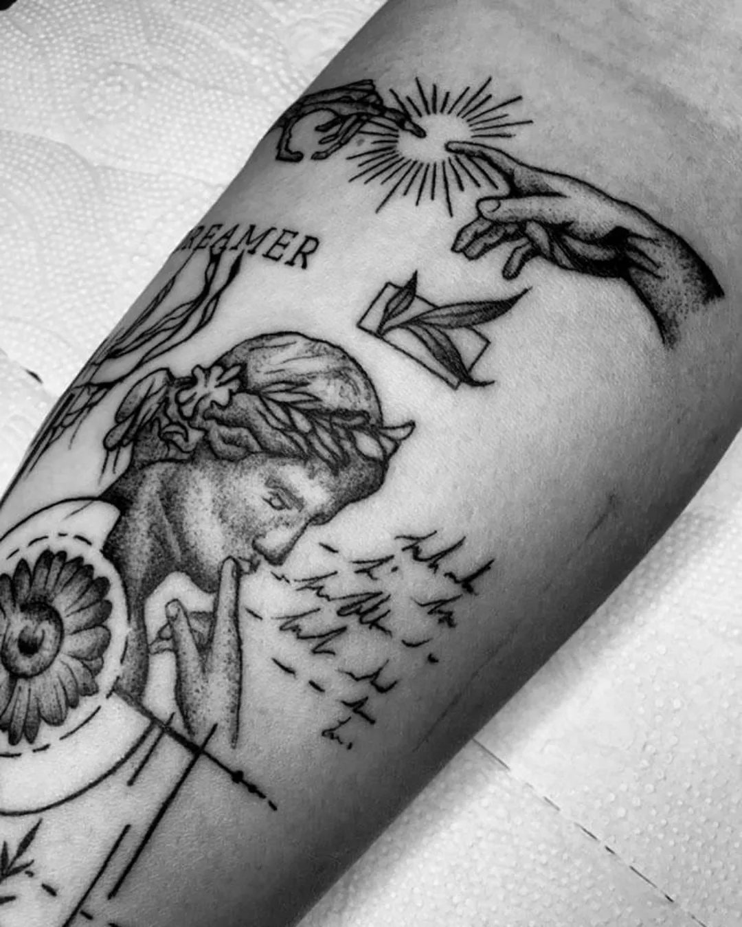 Tatuagem em preto e cinza no antebraço: busto grego, girassol, fases da lua, mãos da Criação de Adão e pássaros, com a palavra Dreamer