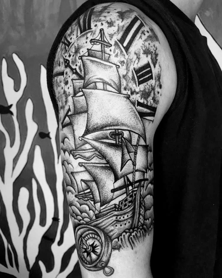 Tatuagem ilustrativa em preto e cinza no braço: navio a vela, relógio de bolso e bússola