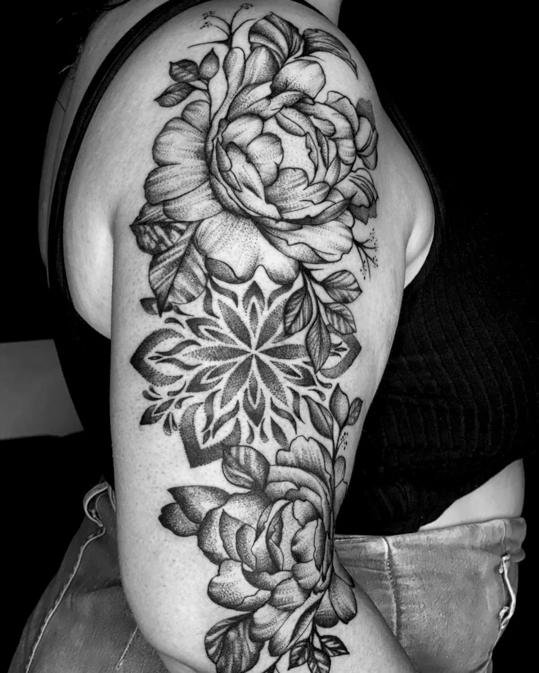 Tatuagem em pontilhismo no braço: peônias em volta de uma mandala ornamental, em preto e cinza